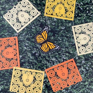Puede incluir: Una cadena de recortes de papel amarillo y naranja con un dise&ntilde;o de coraz&oacute;n, colgando sobre un fondo verde de hojas. Una mariposa est&aacute; en el centro de la cadena.