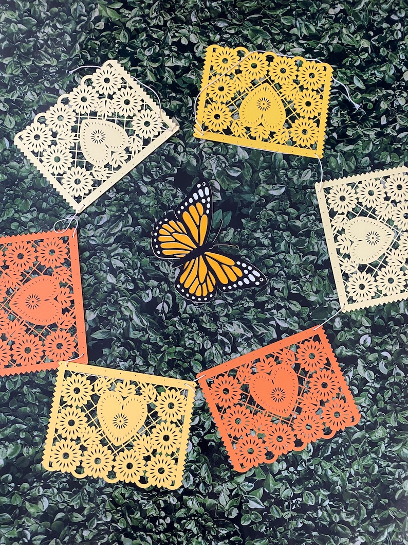 Puede incluir: Una cadena de recortes de papel amarillo y naranja con un dise&ntilde;o de coraz&oacute;n, colgando sobre un fondo verde de hojas. Una mariposa est&aacute; en el centro de la cadena.