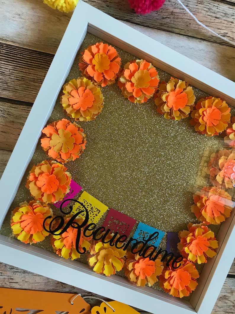 May include: A white shadow box with a gold glitter background. The box is decorated with orange paper flowers and a black paper banner that says "Recuerdame".