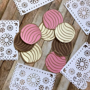 May include: A white paper garland with a scalloped edge and a repeating pattern of flowers and suns. The garland is strung with brown, pink, and white paper circles with a swirl pattern.