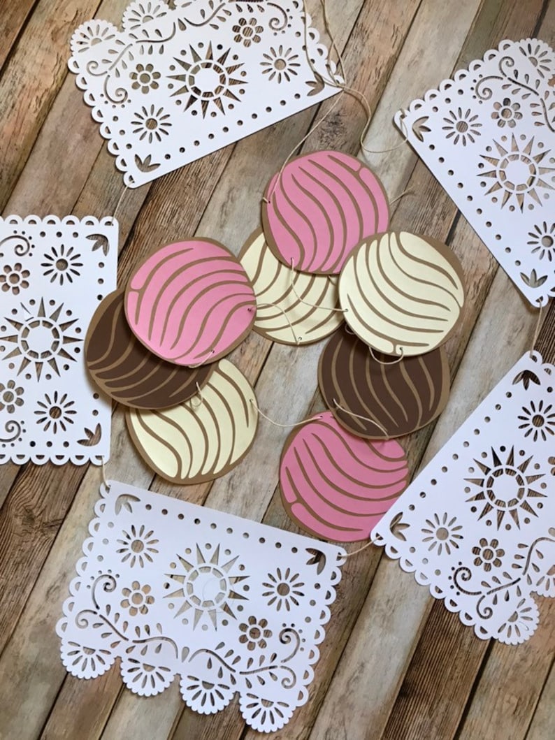 May include: A white paper garland with a scalloped edge and a repeating pattern of flowers and suns. The garland is strung with brown, pink, and white paper circles with a swirl pattern.