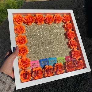 May include: A white picture frame with a gold glitter background and orange paper flowers around the border. The frame has a clear plastic front and the text "Amor eterno e inrvidable" is visible inside the frame.