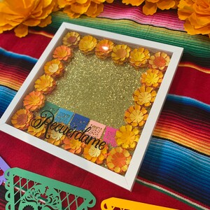 May include: A white picture frame with a gold glitter background and paper flowers in shades of orange and yellow. The frame is decorated with colorful paper flags that say "Recuerdame".