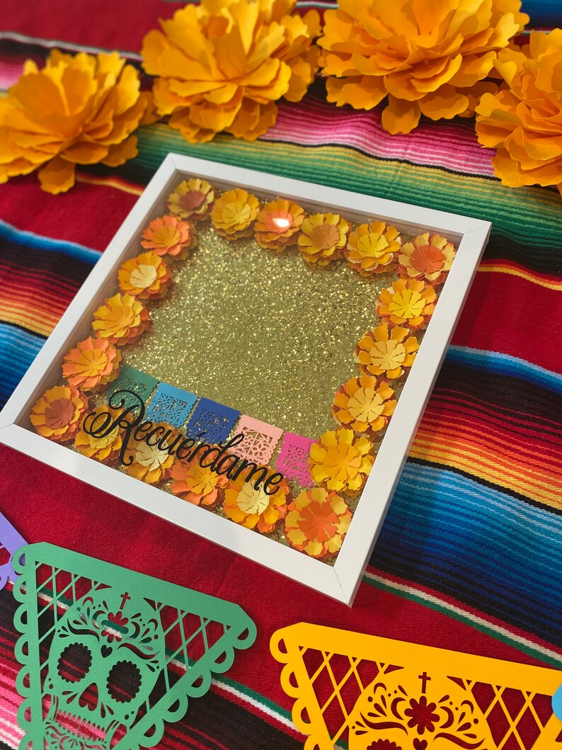 May include: A white picture frame with a gold glitter background and paper flowers in shades of orange and yellow. The frame is decorated with colorful paper flags that say "Recuerdame".