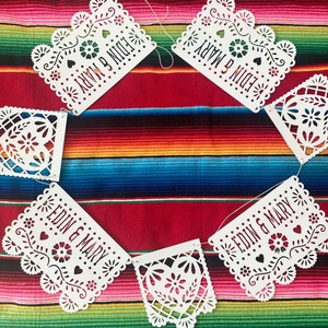 May include: White paper banner with cut-out designs, strung together with white string. Each banner has a heart shape with a floral design and the text "EDIN & MARY" written on it.