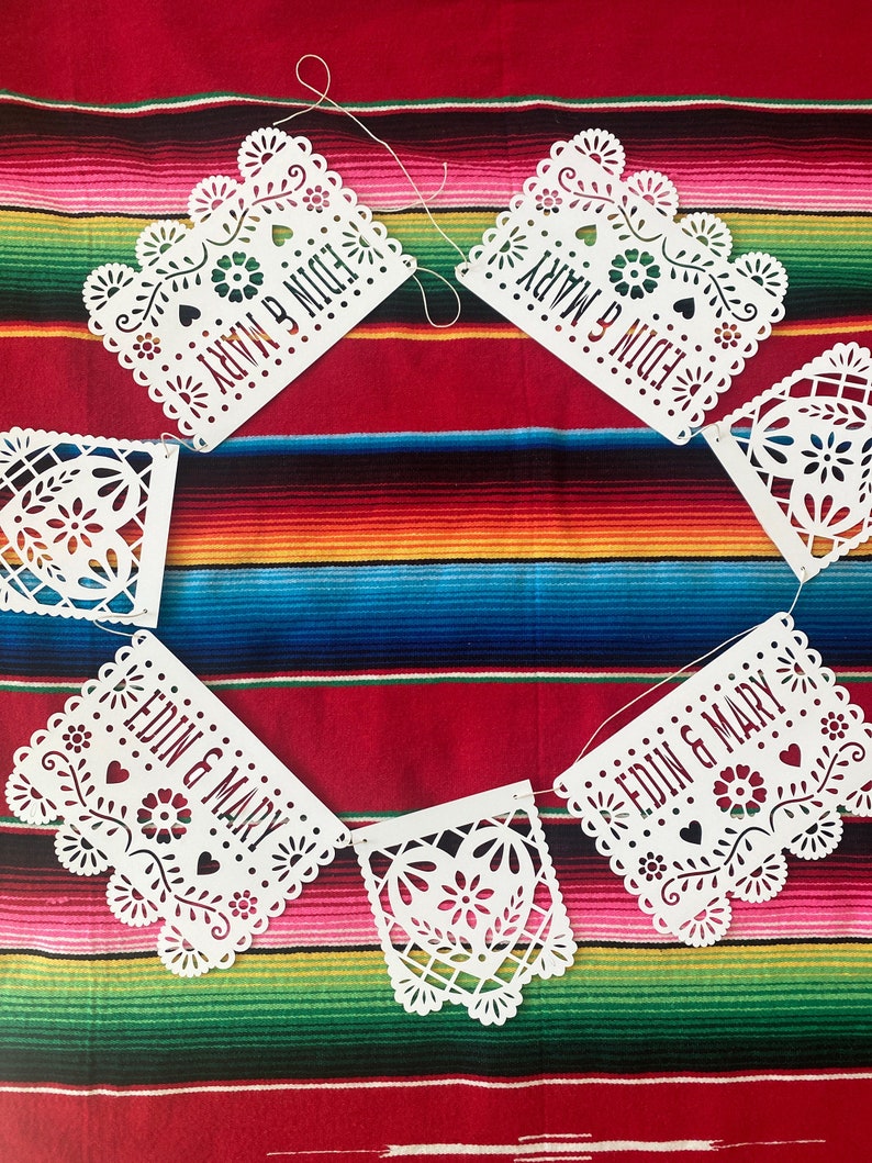 May include: White paper banner with cut-out designs, strung together with white string. Each banner has a heart shape with a floral design and the text "EDIN & MARY" written on it.