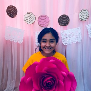 May include: A young person wearing a yellow shirt holds a large, bright pink paper flower in front of a pink and white backdrop. The backdrop has a garland of paper circles with brown, white, and pink stripes.