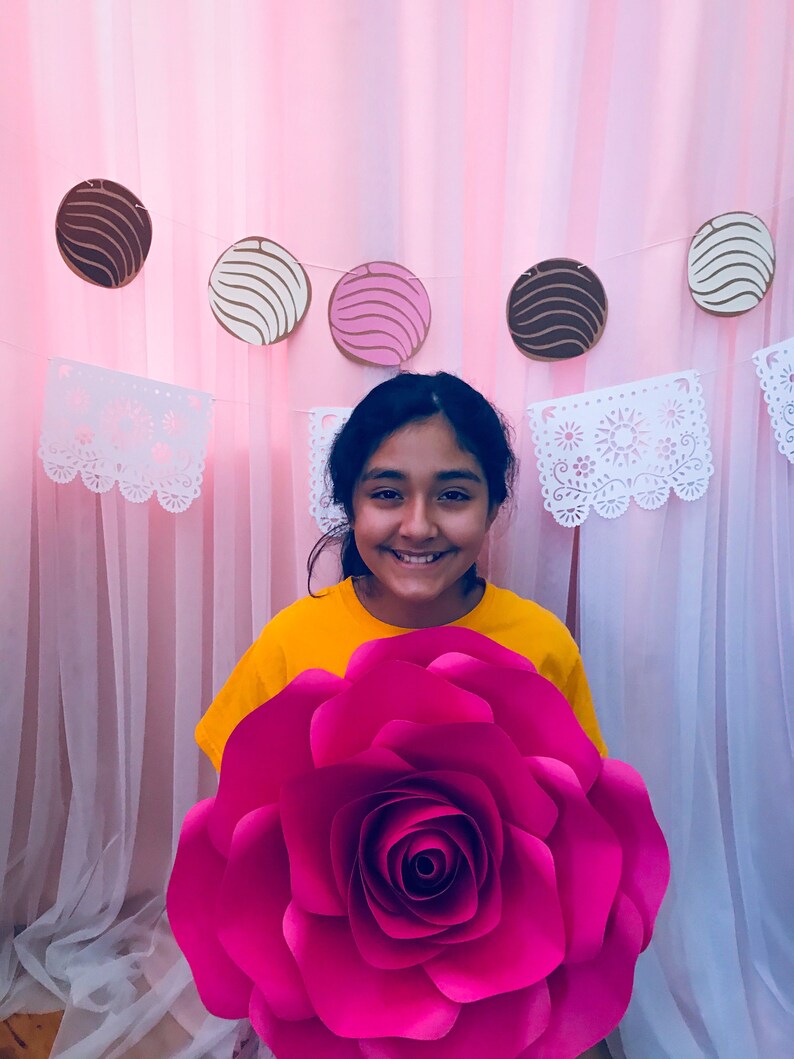 May include: A young person wearing a yellow shirt holds a large, bright pink paper flower in front of a pink and white backdrop. The backdrop has a garland of paper circles with brown, white, and pink stripes.