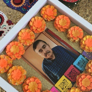 May include: A white picture frame with a photo of a man in a blue plaid shirt. The frame is decorated with orange marigold flowers and colorful paper banners. The background is a gold sparkly surface.