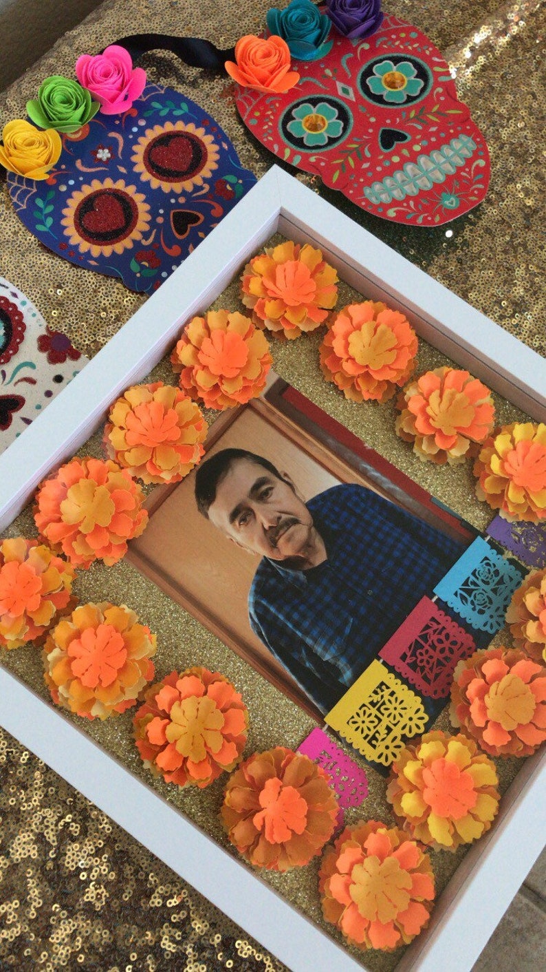 May include: A white picture frame with a photo of a man in a blue plaid shirt. The frame is decorated with orange marigold flowers and colorful paper banners. The background is a gold sparkly surface.