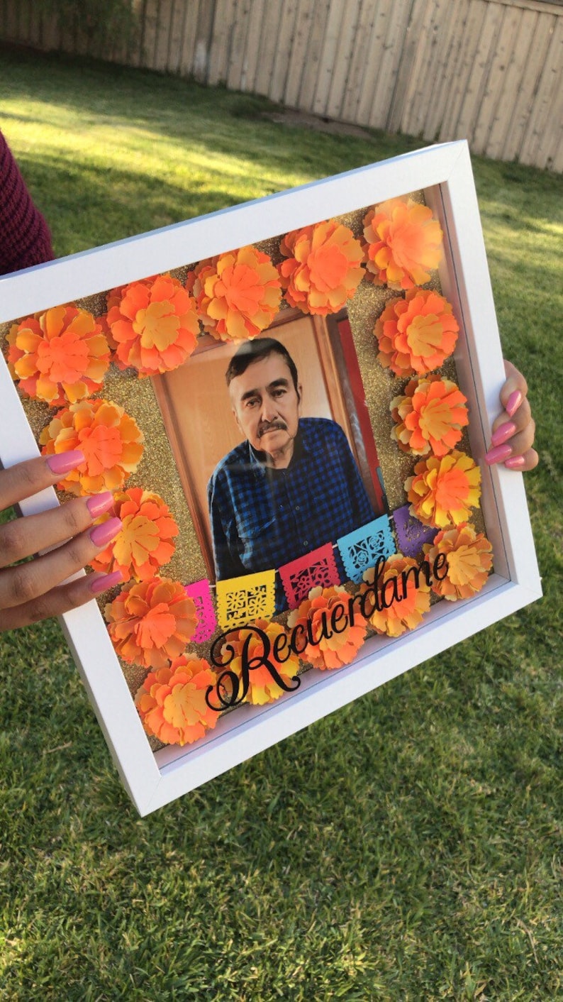 May include: A white shadow box with a photo of a man inside. The box is decorated with orange paper flowers and the words "Recuerdame" in black lettering.