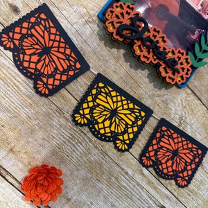 May include: A decorative banner featuring three rectangular black paper cutouts with intricate butterfly and floral patterns in orange and yellow, accompanied by a layered frame with a photograph of a person playing guitar and a small orange paper flower.
