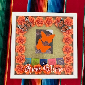May include: A white picture frame with a gold glitter background. The frame is decorated with orange paper flowers and butterflies. The frame contains a photo and the text "Amor Eterno".