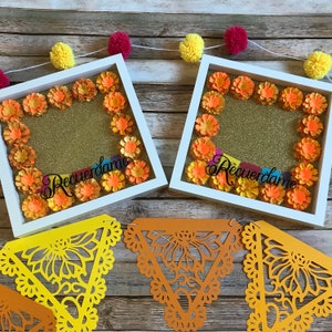 May include: Two white picture frames with gold glitter backgrounds and orange paper flowers. The frames have the Spanish phrase "Recu&eacute;rdame" written in black. The frames are sitting on a wooden surface with a yellow and orange paper garland.