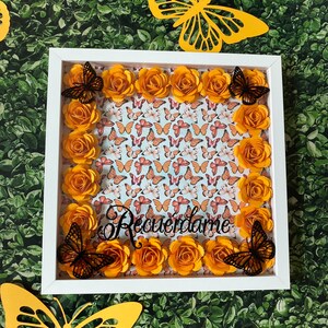 May include: A white picture frame with a white mat and a light blue background with a pattern of orange butterflies. The frame is decorated with orange paper flowers and black butterflies. The text "Recuerdame" is written in black cursive lettering.