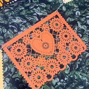 Puede incluir: Bandera de papel naranja con un coraz&oacute;n y dise&ntilde;os florales. La bandera est&aacute; recortada con detalles intrincados y tiene un borde festoneado.