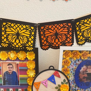 May include: A framed photo with a colorful background and a man wearing a plaid shirt. The frame is decorated with yellow paper flowers. A black and orange paper banner with butterflies hangs above the frame.