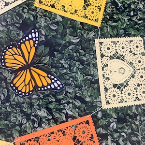 Puede incluir: Una cadena de recortes de papel amarillo y naranja con un dise&ntilde;o de mariposa. Los recortes est&aacute;n unidos con una cuerda blanca y cuelgan sobre un fondo verde de hojas.