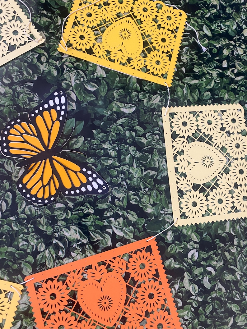 Puede incluir: Una cadena de recortes de papel amarillo y naranja con un dise&ntilde;o de mariposa. Los recortes est&aacute;n unidos con una cuerda blanca y cuelgan sobre un fondo verde de hojas.