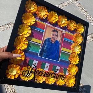 May include: A black picture frame with a photo of a man in a blue plaid shirt. The frame is decorated with yellow paper flowers and the text "Recuérdame" in black cursive lettering.