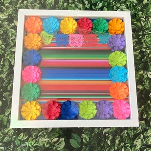 May include: A white frame with a colorful Mexican serape pattern inside. The serape is surrounded by colorful paper flowers in a variety of colors, including red, orange, yellow, green, blue, and purple.