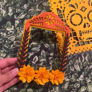 May include: A layered orange and red paper frame with black accents and a scalloped top. The frame is decorated with three orange paper flowers.
