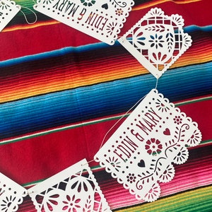 May include: White paper banner with a cut-out design, featuring the words "Edin & Mary" in a red font. The banner is strung together with white thread and is hanging against a colorful striped background.
