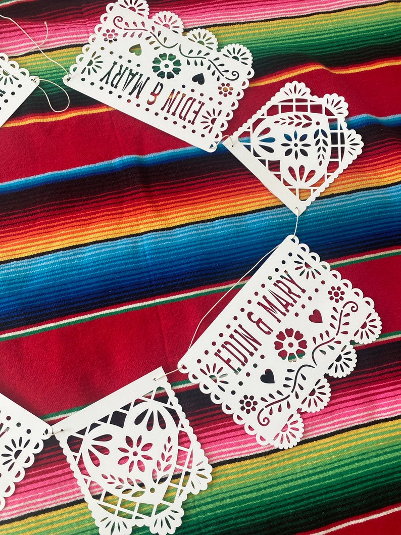 May include: White paper banner with a cut-out design, featuring the words "Edin & Mary" in a red font. The banner is strung together with white thread and is hanging against a colorful striped background.