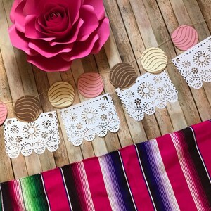May include: A festive garland with pink, brown, and yellow paper circles, white paper doilies, and a pink paper flower. The garland is hanging on a wooden background. A pink and green striped blanket is in the foreground.