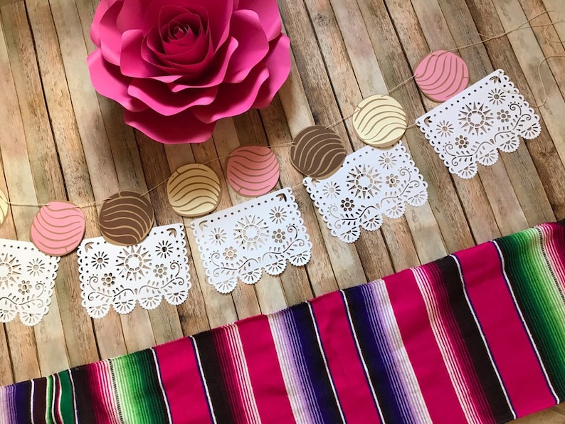 May include: A festive garland with pink, brown, and yellow paper circles, white paper doilies, and a pink paper flower. The garland is hanging on a wooden background. A pink and green striped blanket is in the foreground.