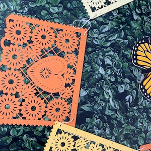 Puede incluir: Una cadena de recortes de papel naranja y amarillo con un dise&ntilde;o floral. Los recortes est&aacute;n unidos con una cuerda blanca y cuelgan sobre un fondo verde.