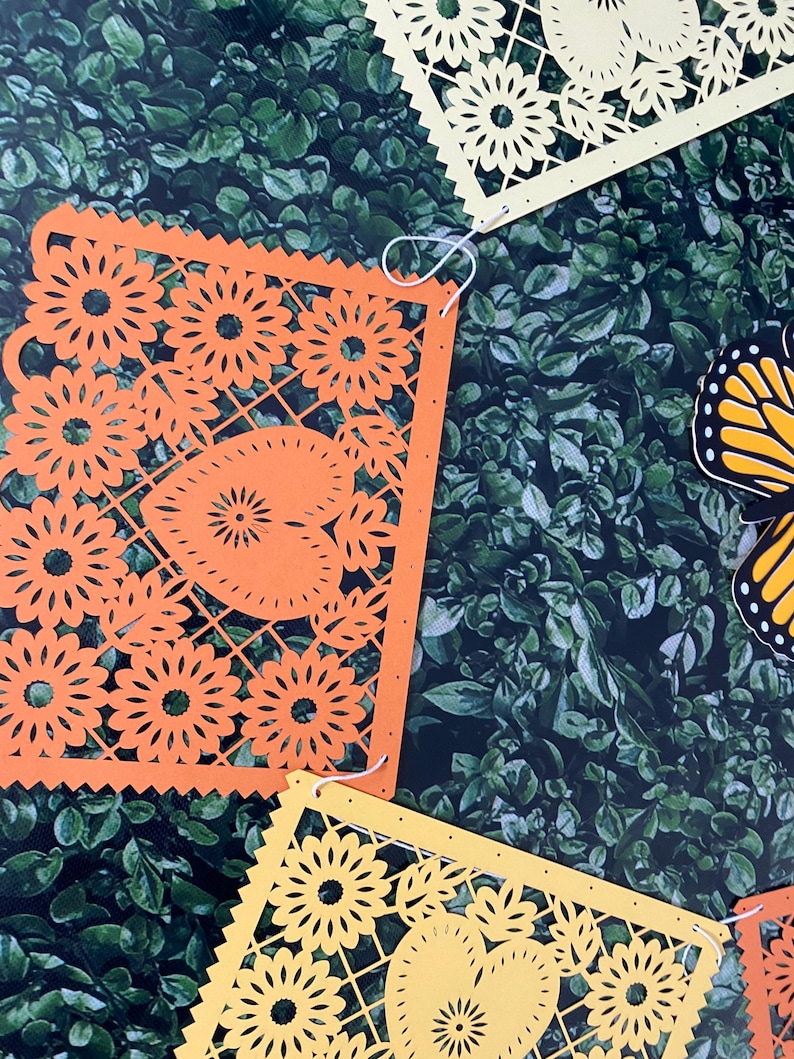 Puede incluir: Una cadena de recortes de papel naranja y amarillo con un dise&ntilde;o floral. Los recortes est&aacute;n unidos con una cuerda blanca y cuelgan sobre un fondo verde.