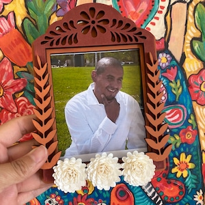 Puede incluir: Un marco de fotos de madera con un diseño tallado y flores blancas. El marco contiene una foto de una persona sonriendo. El marco está colocado sobre un fondo colorido.