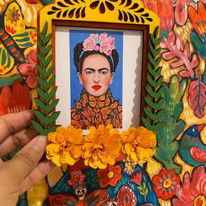 May include: A yellow wooden frame with a cutout design, featuring a picture of Frida Kahlo with a butterfly headdress. The frame is decorated with green leaves and yellow paper flowers.