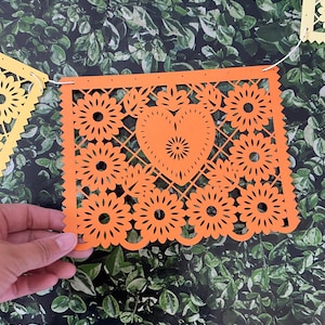 Puede incluir: Bandera de papel naranja con un coraz&oacute;n y un dise&ntilde;o floral. La bandera est&aacute; enhebrada con hilo blanco y tiene agujeros perforados en los bordes.