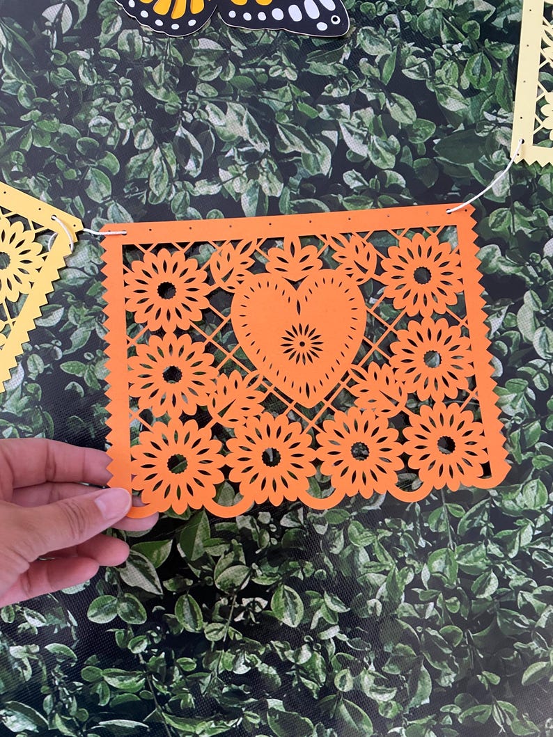 Puede incluir: Bandera de papel naranja con un coraz&oacute;n y un dise&ntilde;o floral. La bandera est&aacute; enhebrada con hilo blanco y tiene agujeros perforados en los bordes.