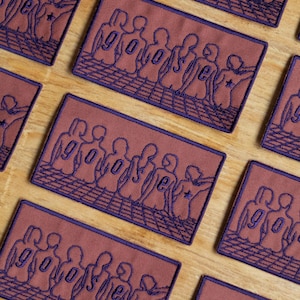 May include: Rectangular embroidered patches in a repeating pattern. Each patch features a maroon background with a purple outline of figures and the word "go" and the letter "e".