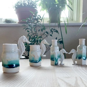 Puede incluir: Cinco figuritas de caballito de mar de cerámica con un esmalte azul verdoso y blanco. Los caballitos de mar están dispuestos en fila sobre una superficie de madera.