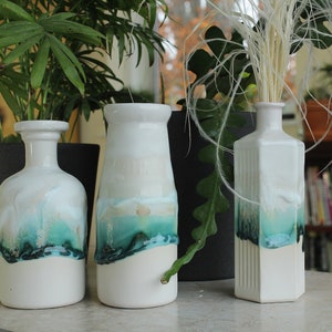 May include: Three white ceramic vases with a teal and white glaze. The vases are different shapes and sizes. The vase on the right has a rectangular shape with vertical lines. The vase in the middle is a rounder shape. The vase on the left is a more traditional bottle shape.