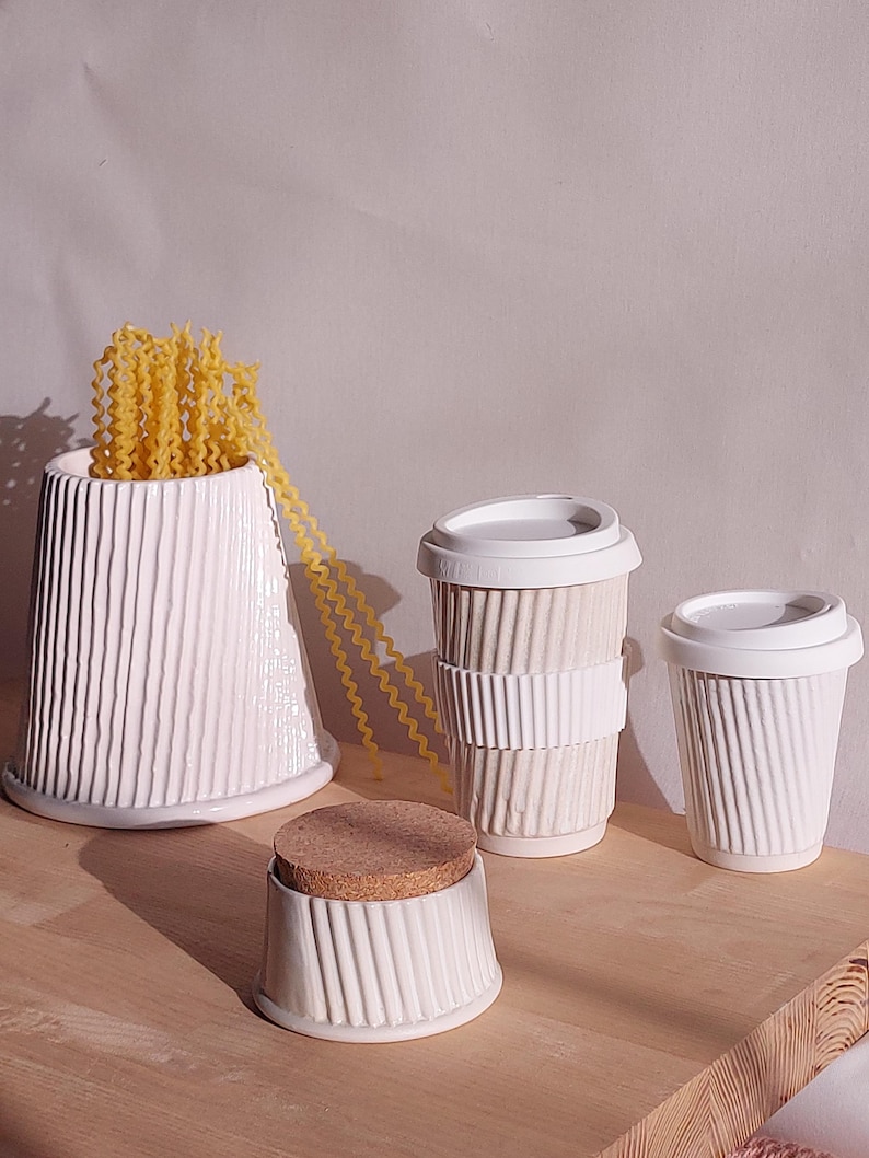 May include: A collection of white ceramic kitchenware and a pasta-filled vase. The items include a vase with vertical ridges, two coffee cups with lids, and a small container with a cork lid. The pasta is yellow and the items are on a wooden surface.