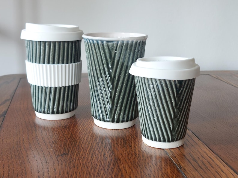 May include: Three ceramic travel mugs with textured, dark green exteriors and white accents. The mugs vary in size, with one featuring a white ribbed sleeve and another with a white lid. The mugs are on a wooden surface.