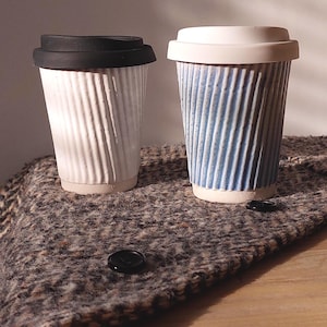 May include: Two ceramic travel mugs with ribbed sides and lids. One mug is white with a black lid, the other is blue and white with a white lid. The mugs are on a textured, brown and gray surface.
