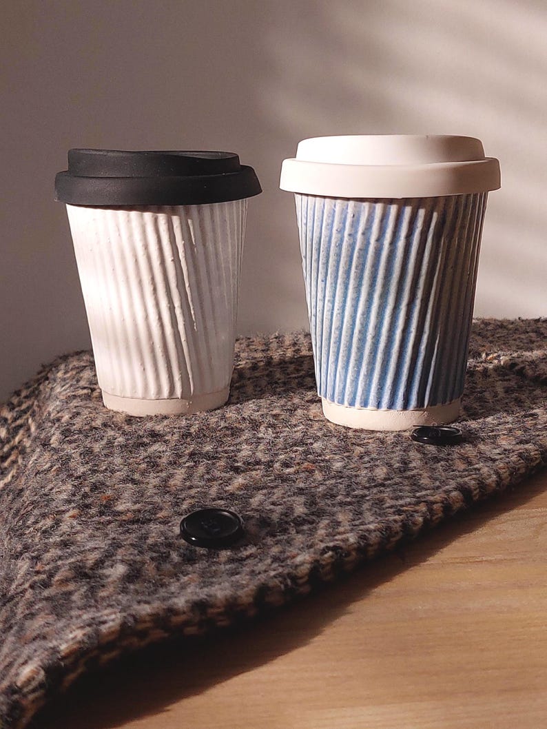 May include: Two ceramic travel mugs with ribbed sides and lids. One mug is white with a black lid, the other is blue and white with a white lid. The mugs are on a textured, brown and gray surface.