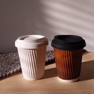 May include: Two ceramic travel mugs with lids. One is off-white with a white lid, the other is brown with a black lid. Both mugs have a ridged design. The mugs are on a wooden surface.