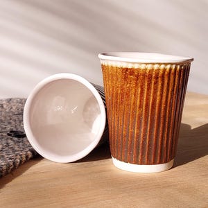 May include: Two brown paper coffee cups with a ribbed texture, one upright and the other lying on its side. The cups have a white rim and base. The cups are on a wooden surface.