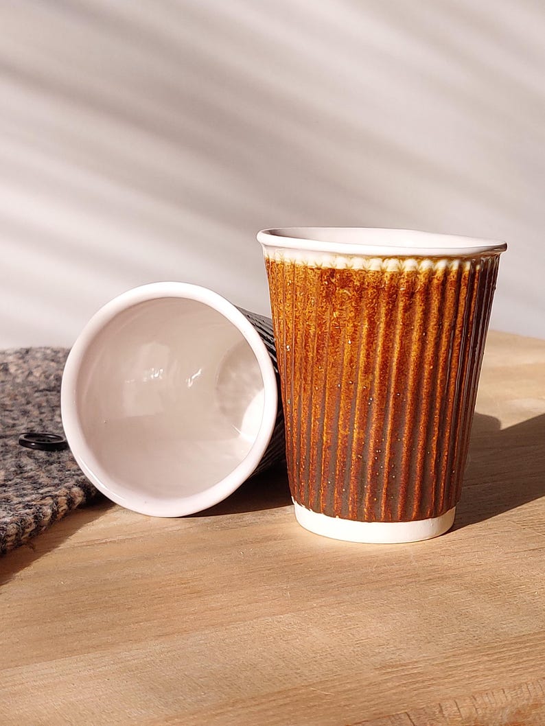 May include: Two brown paper coffee cups with a ribbed texture, one upright and the other lying on its side. The cups have a white rim and base. The cups are on a wooden surface.