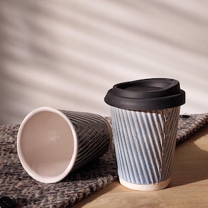 May include: Two ceramic travel mugs. One is blue with a black lid, the other is black. Both have a ridged, textured design. The mugs are on a textured, gray surface.