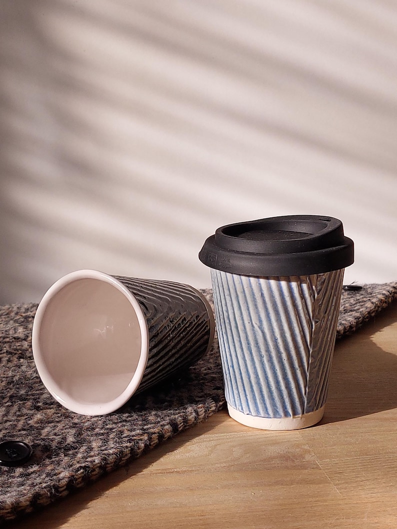 May include: Two ceramic travel mugs. One is blue with a black lid, the other is black. Both have a ridged, textured design. The mugs are on a textured, gray surface.