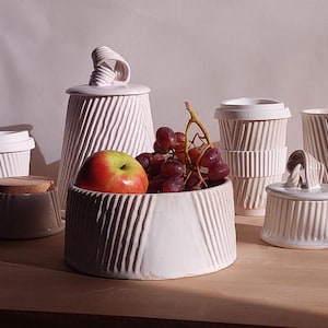 May include: A collection of white ceramic kitchenware with a textured, ribbed design. Includes a bowl with an apple and grapes, a lidded jar, a small container with a cork lid, and several cups. The items are arranged on a wooden surface.