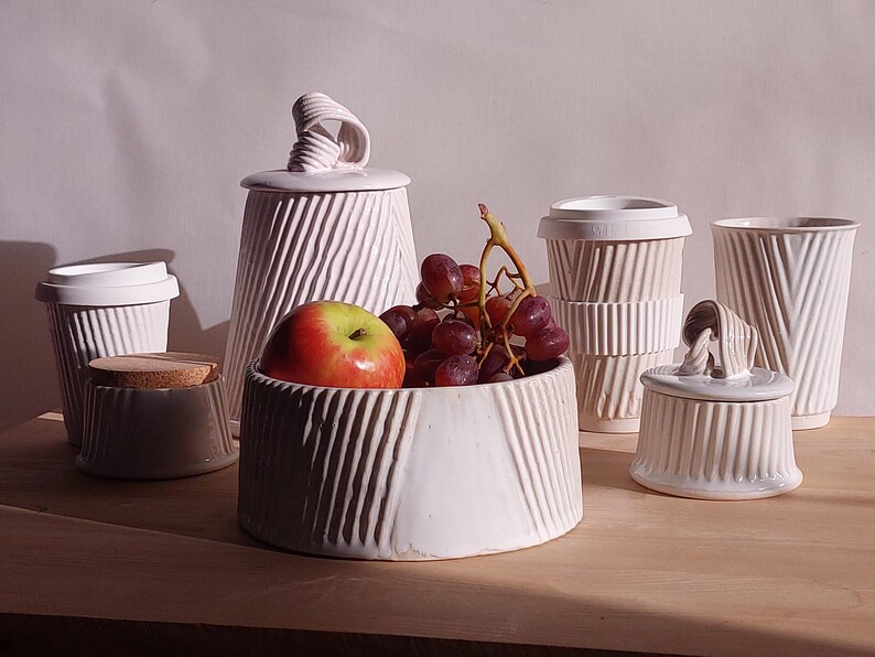 May include: A collection of white ceramic kitchenware with a textured, ribbed design. Includes a bowl with an apple and grapes, a lidded jar, a small container with a cork lid, and several cups. The items are arranged on a wooden surface.